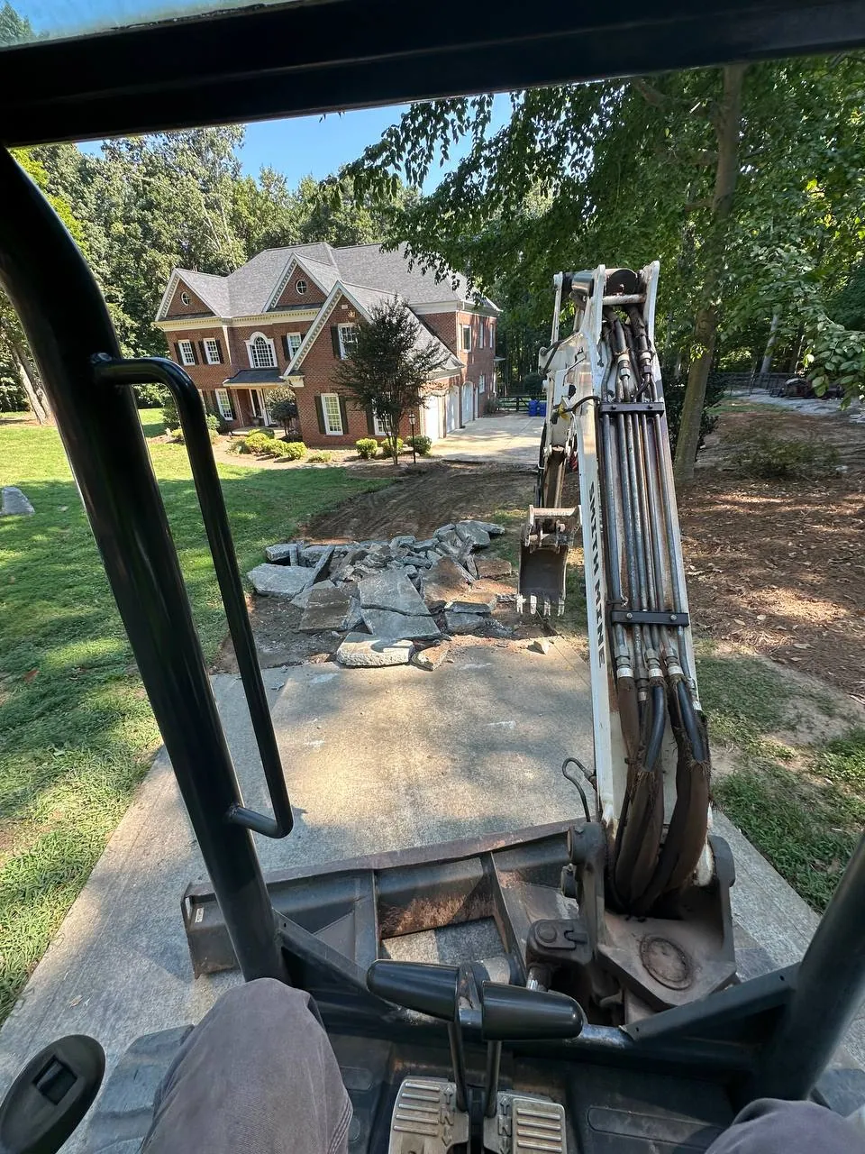 View from the mini excavator cab: SMITH MINE on the boom, old concrete driveway broken into slabs in front of a large brick house