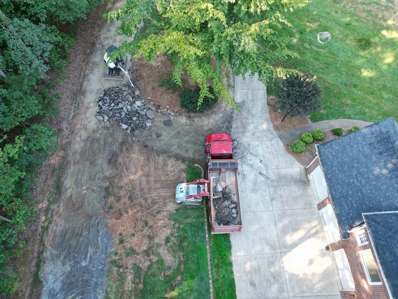 Drone view straight down: Takeuchi track loader dumping broken driveway concrete into a red tandem dump truck beside a brick house