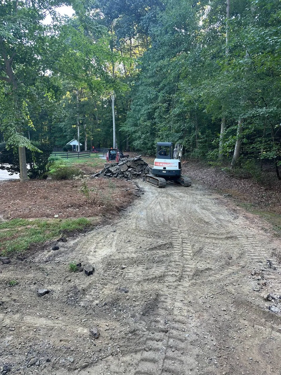 Old driveway torn out and graded flat through the woods, Bobcat mini excavator beside the broken concrete pile, track loader beyond