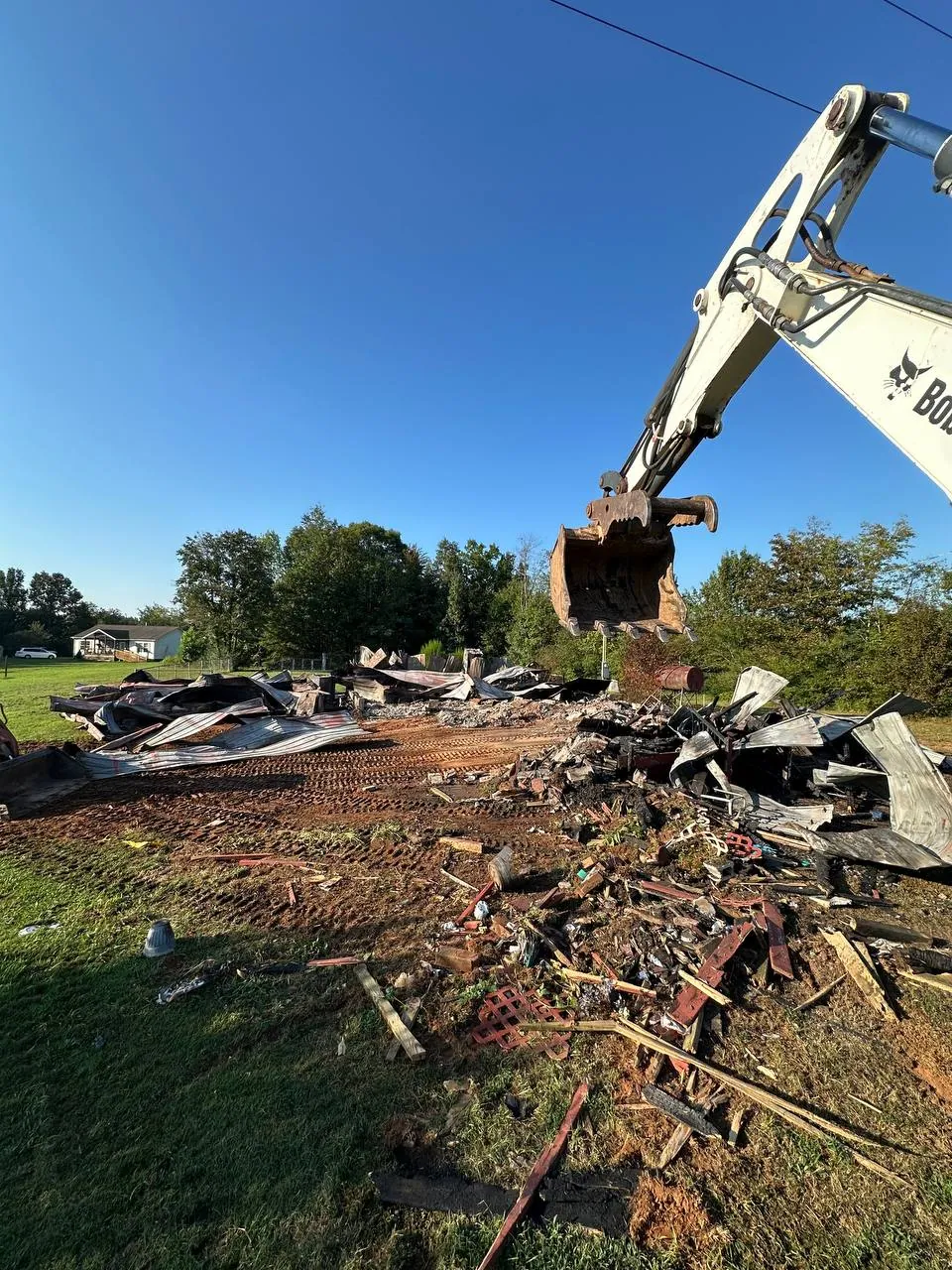 Bobcat mini excavator bucket raised over a burned structure torn down to charred framing and twisted metal roofing
