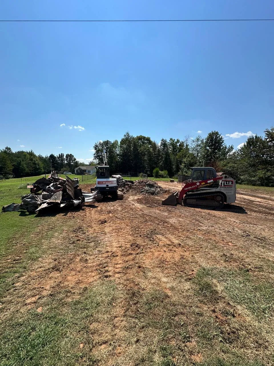 Takeuchi TL12 track loader and Bobcat mini excavator on a scraped red clay lot, torn down metal roofing and debris piled beside them