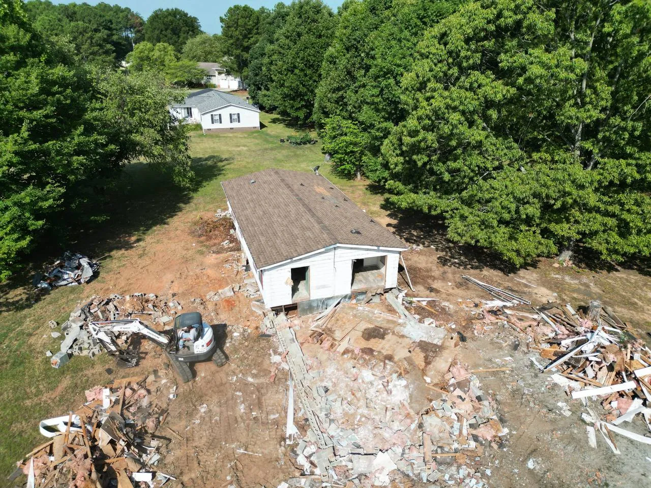 Drone aerial: Bobcat mini excavator tearing down a mobile home under the trees, block, framing and insulation piled around it